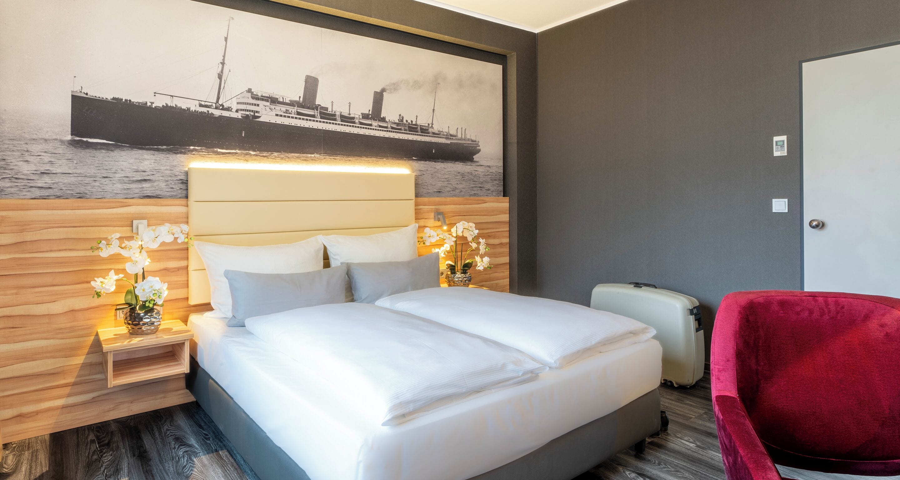 Ein Hotelzimmer mit einem großen Bett, einem roten Sessel, einem Koffer und einem an der Wand hängenden Bild von einem Schiff.