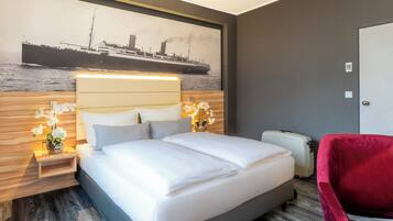Ein Hotelzimmer mit einem großen Bett, einem roten Sessel, einem Koffer und einem an der Wand hängenden Bild von einem Schiff.