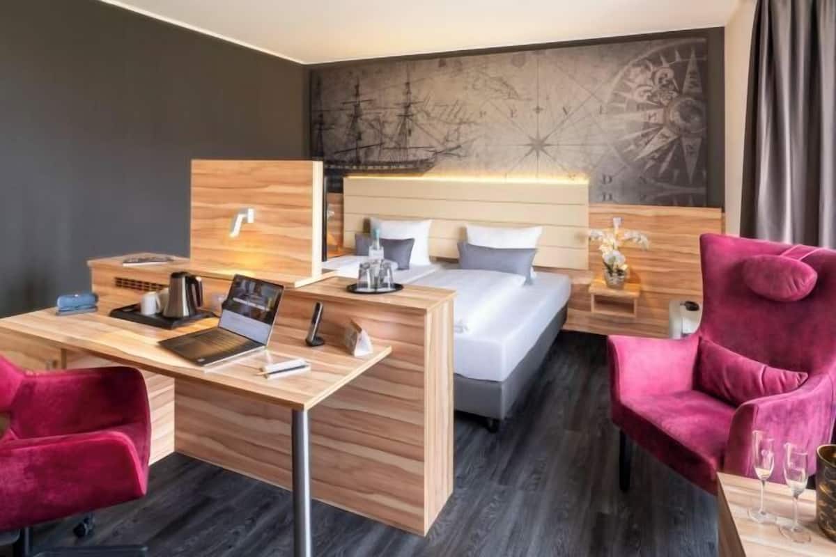 A modern hotel room with a wooden desk, a bed with white bedding, a red chair, and a wall mural.