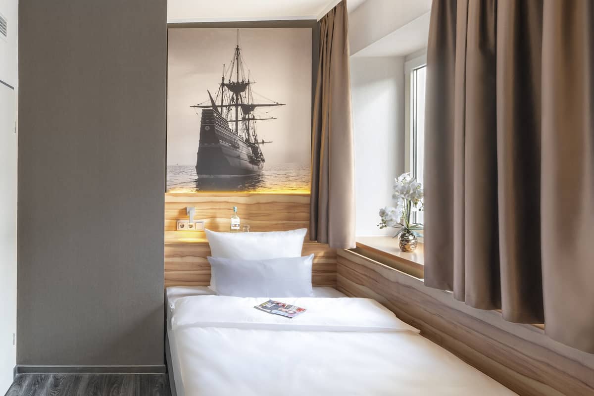 Camera d'albergo con un letto, una finestra con tende, una testiera in legno e un'immagine di una nave appesa al muro.