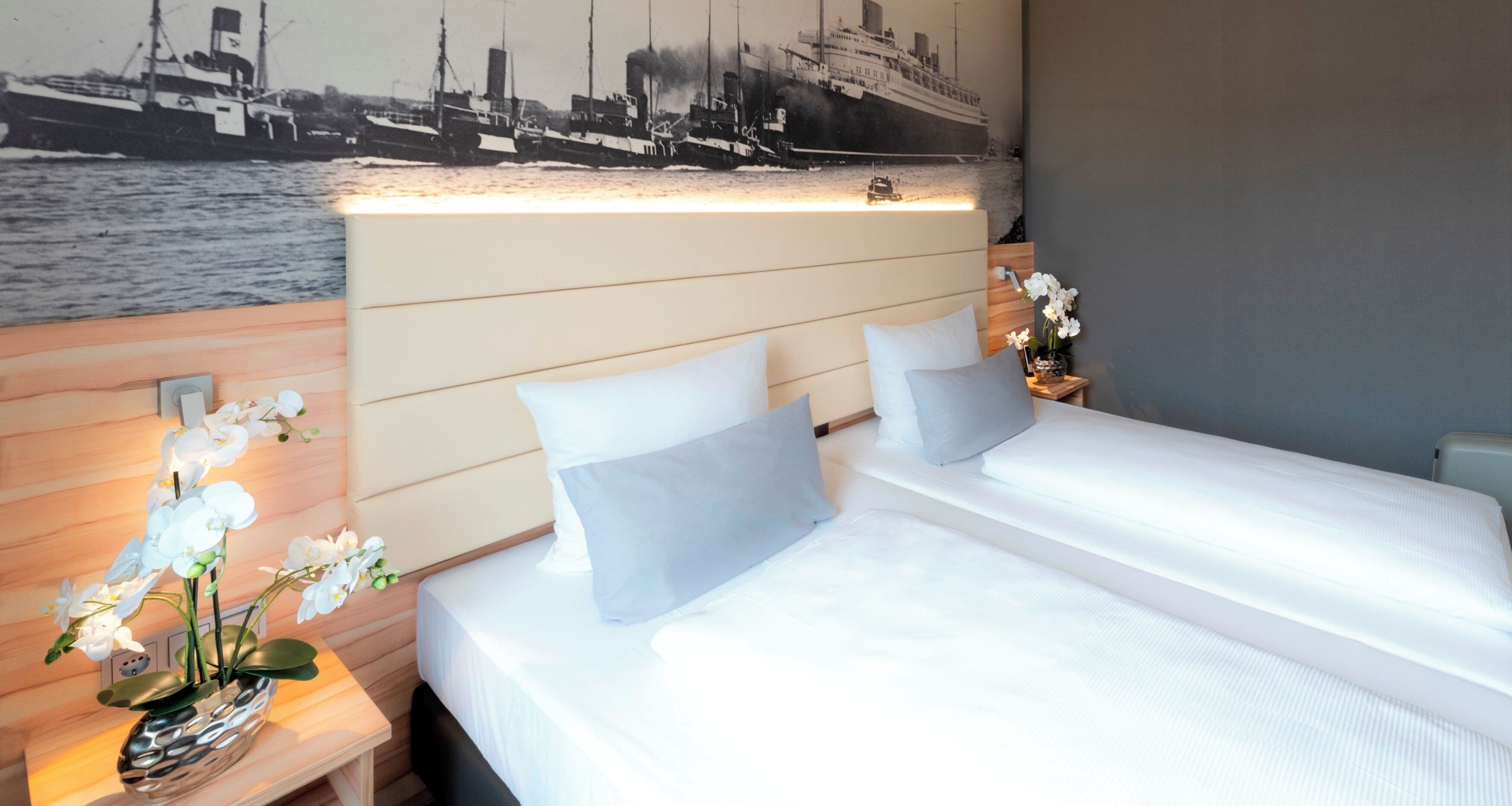 A hotel room with two beds, a headboard with a cityscape mural, a nightstand with a vase of flowers, and a wall-mounted lamp.