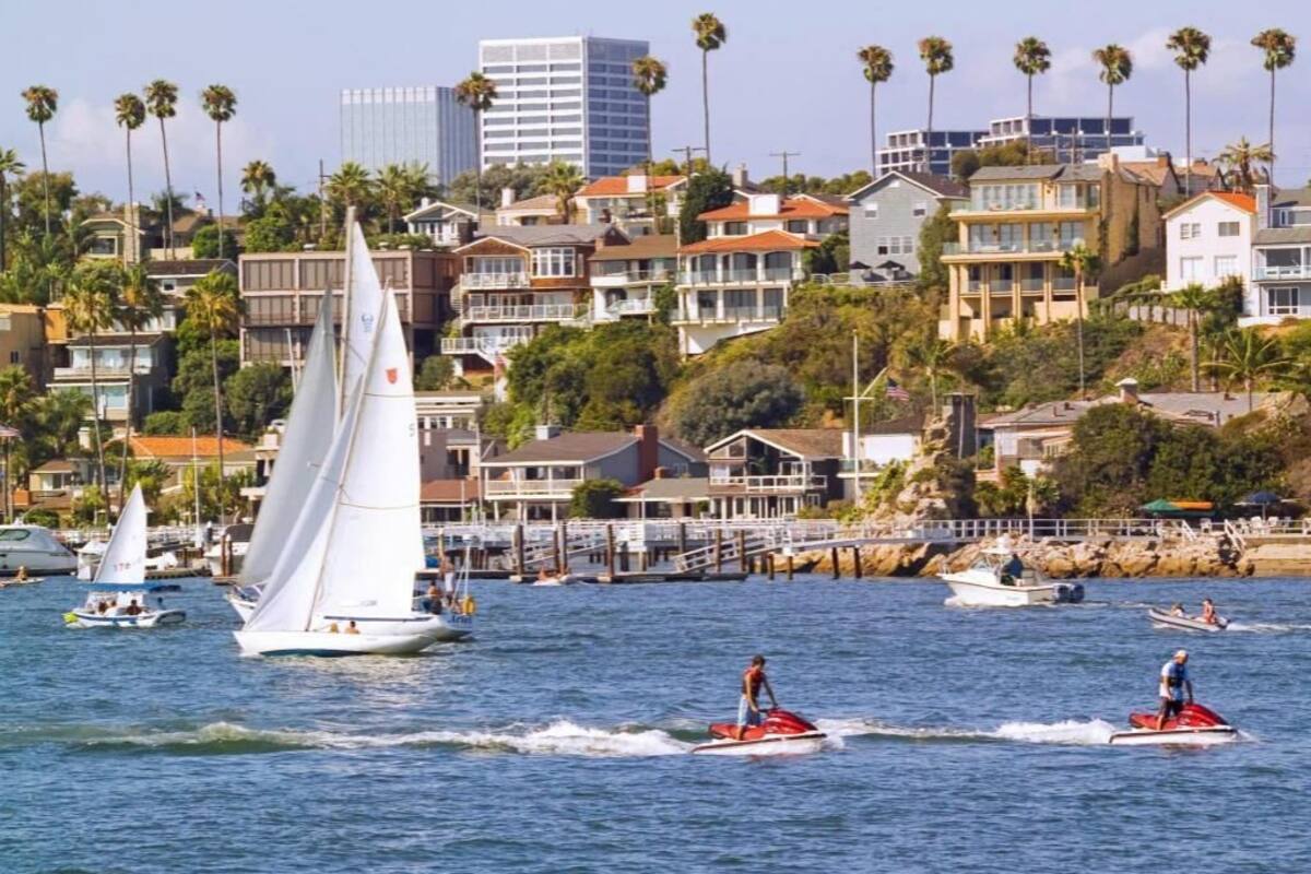 Un paysage urbain côtier avec des voiliers et des jet-skis sur l’eau, des immeubles résidentiels et des palmiers.
