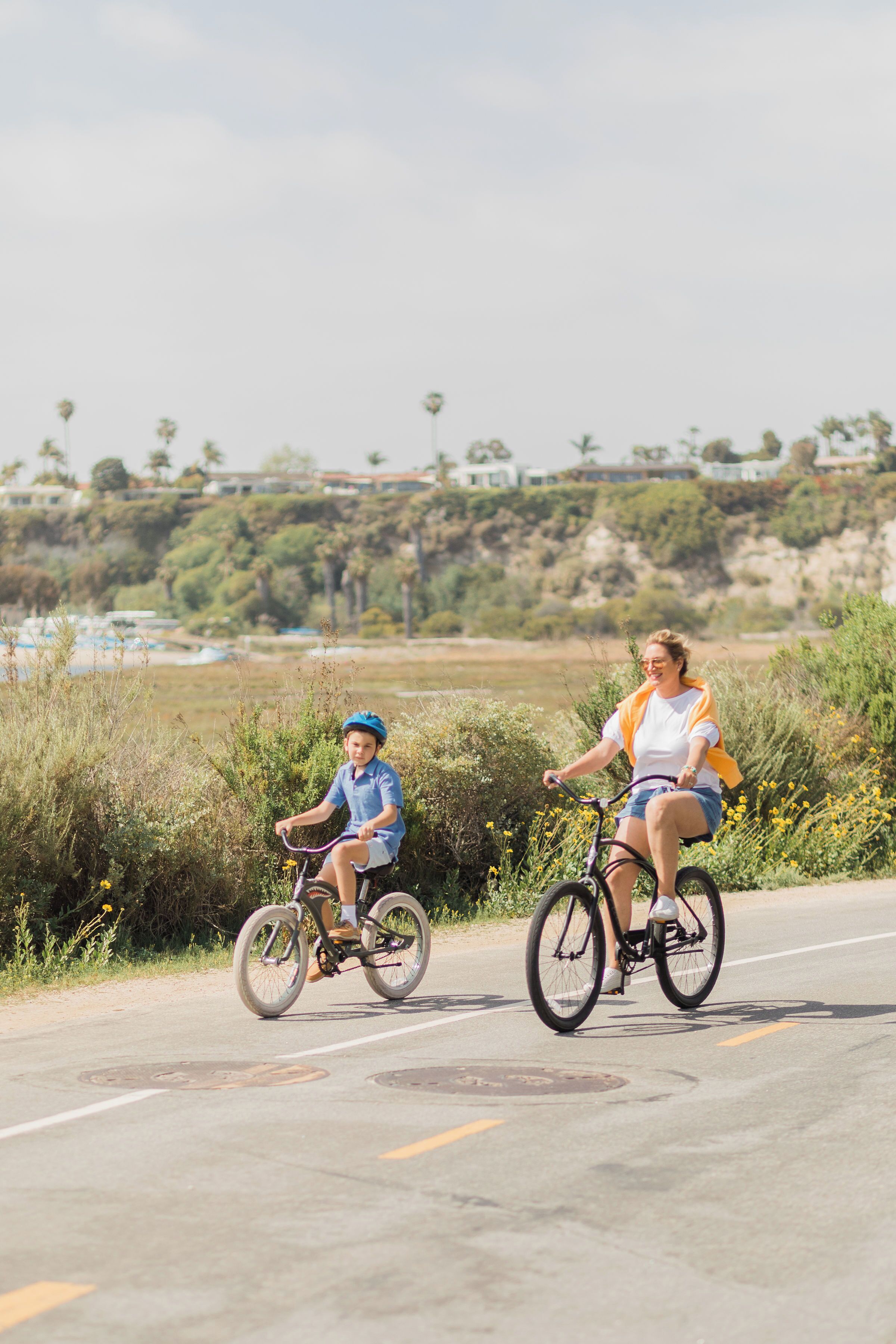 Two people riding bicycles on a road with greenery and buildings in the background.