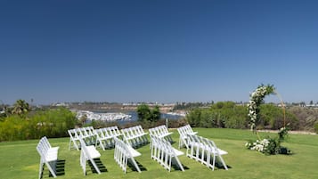 Decoración para una ceremonia de boda con sillas blancas dispuestas sobre un césped, con vista a un cuerpo de agua con barcos y una costa lejana.