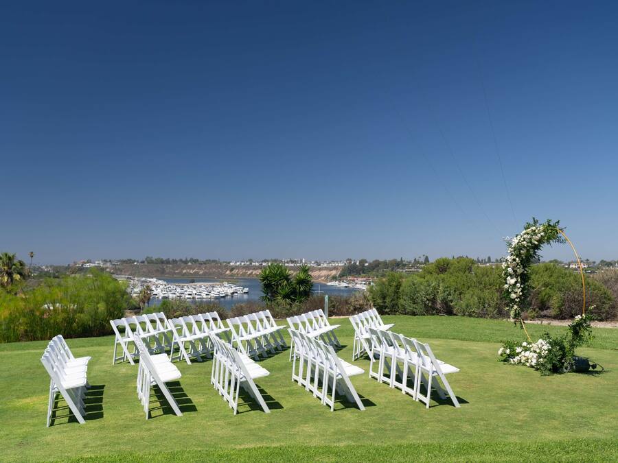 Decoración para una ceremonia de boda con sillas blancas dispuestas sobre un césped, con vista a un cuerpo de agua con barcos y una costa lejana.