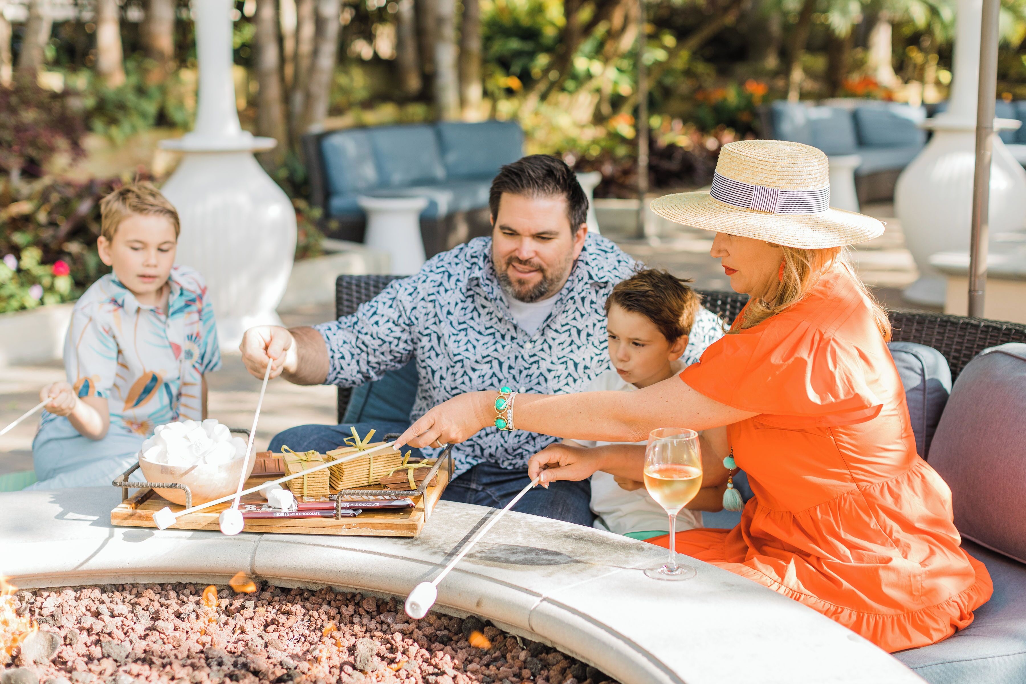 Una familia disfruta de una parrillada con malvaviscos y brochetas, sentada en muebles de exterior con vistas a un jardín.