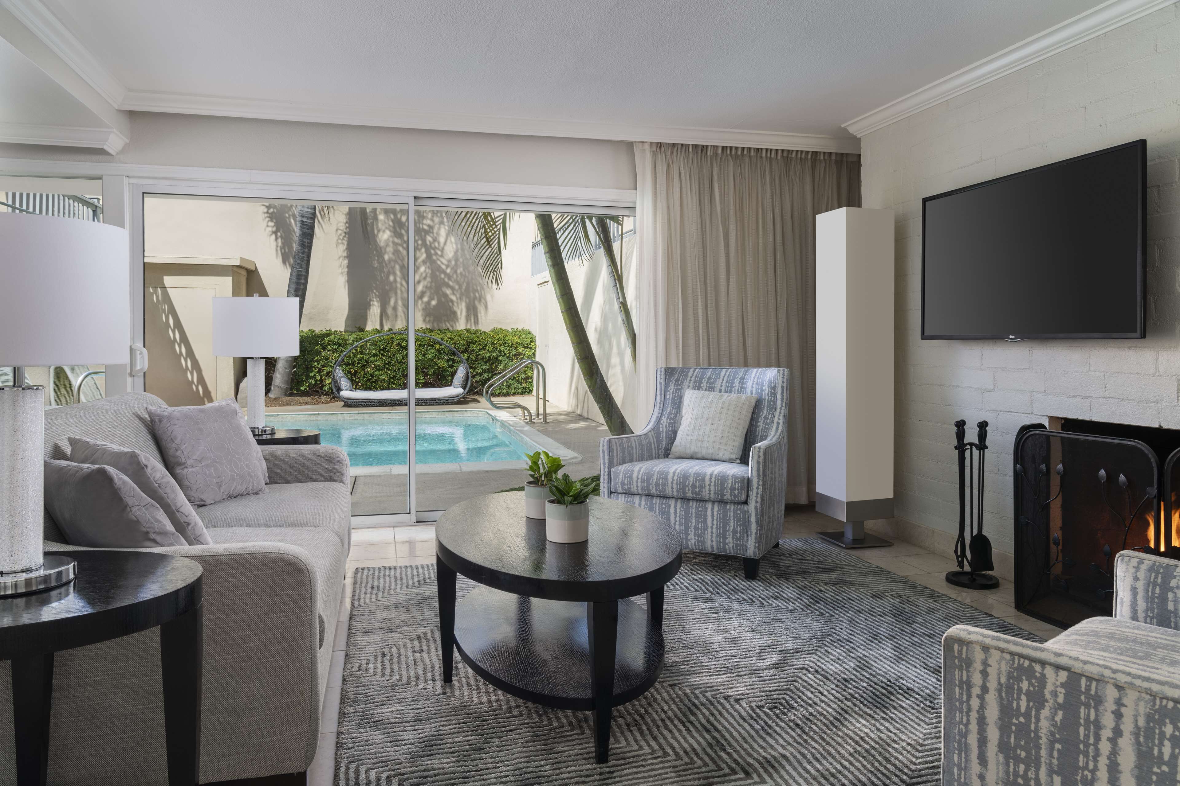 A modern hotel room with a sofa, armchair, coffee table, and a flat-screen TV. A fireplace and a pool are visible through the glass doors.