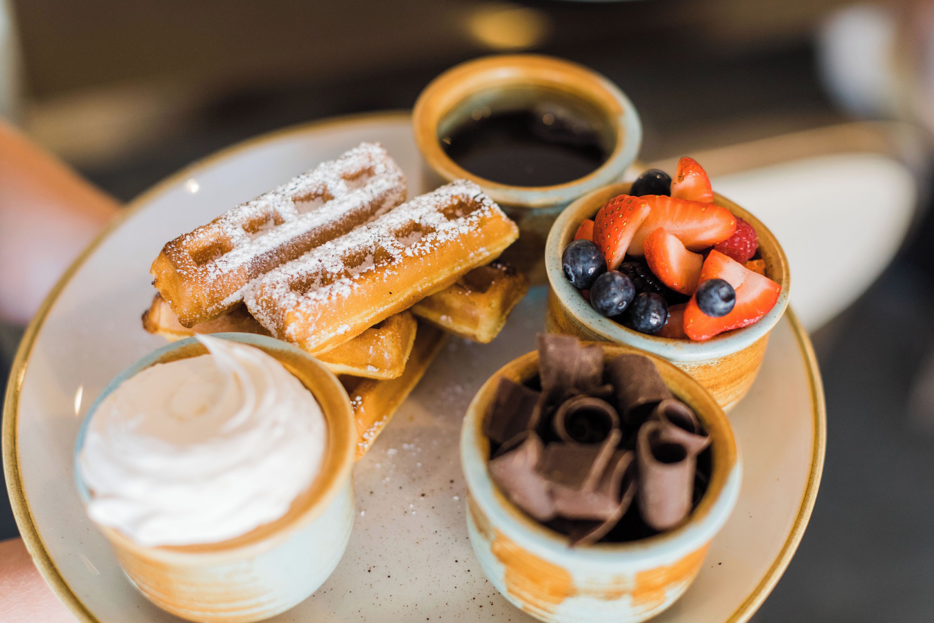 Ein Teller mit Waffeln, eine Schale Erdbeeren, eine Schale Schokolade und eine Schale Schlagsahne.