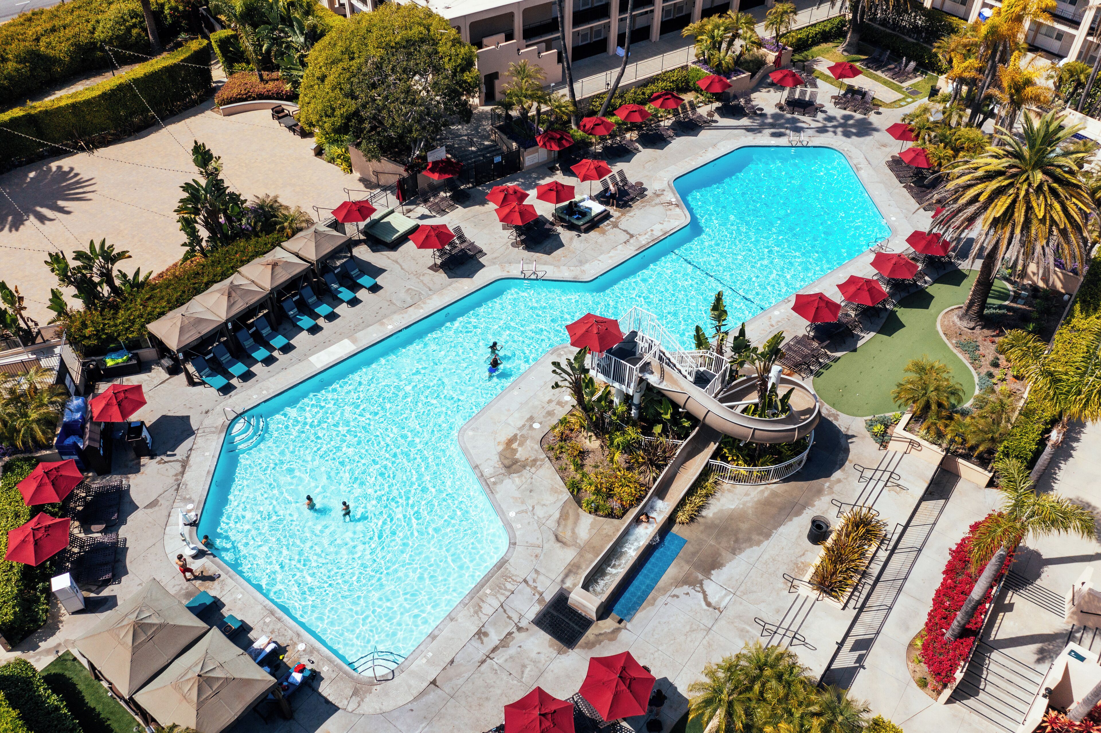 An aerial view of a hotel pool area with multiple swimming pools, sun loungers, and red umbrellas.