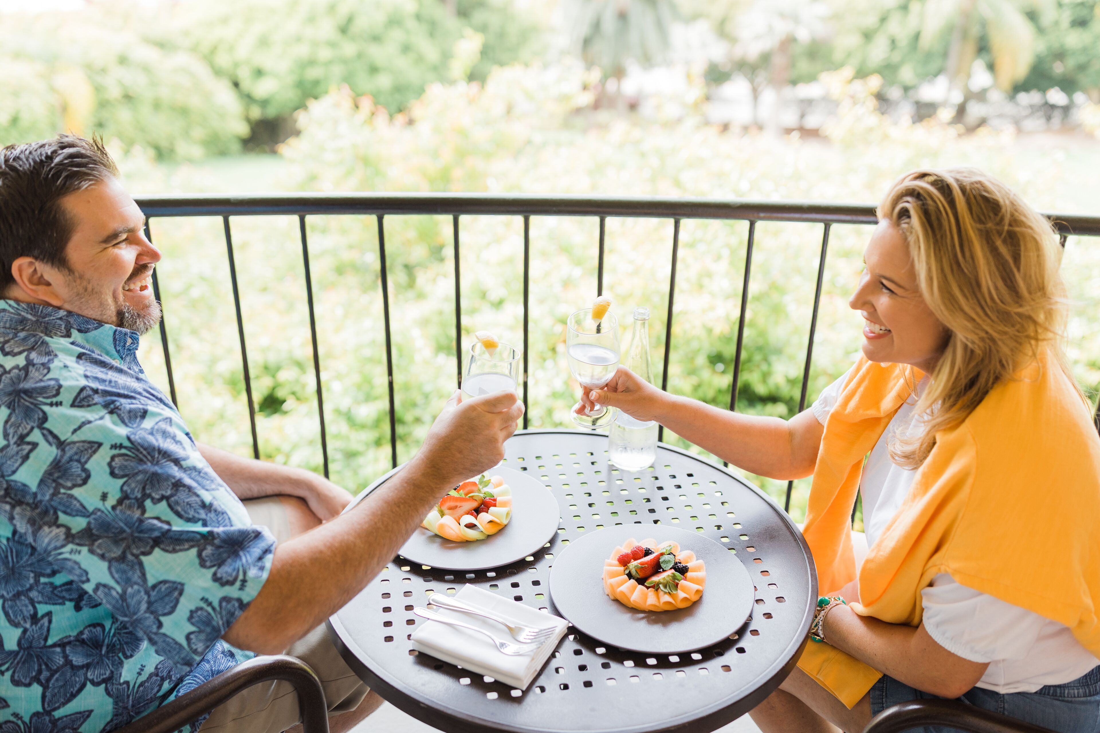 Two people seated at a round table with a garden view, holding drinks and toasting.