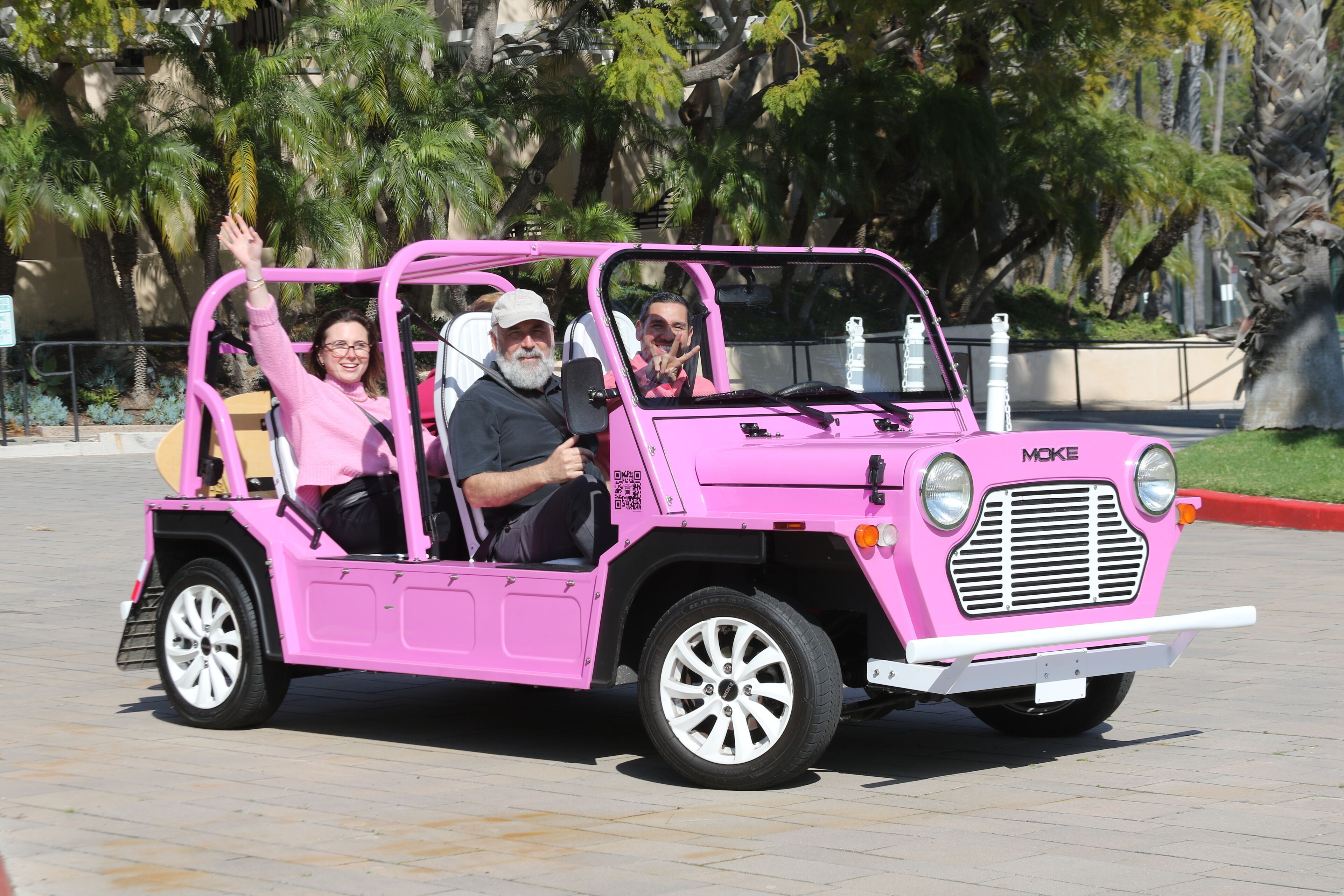 A pink vehicle with passengers, parked on a paved area with trees in the background.