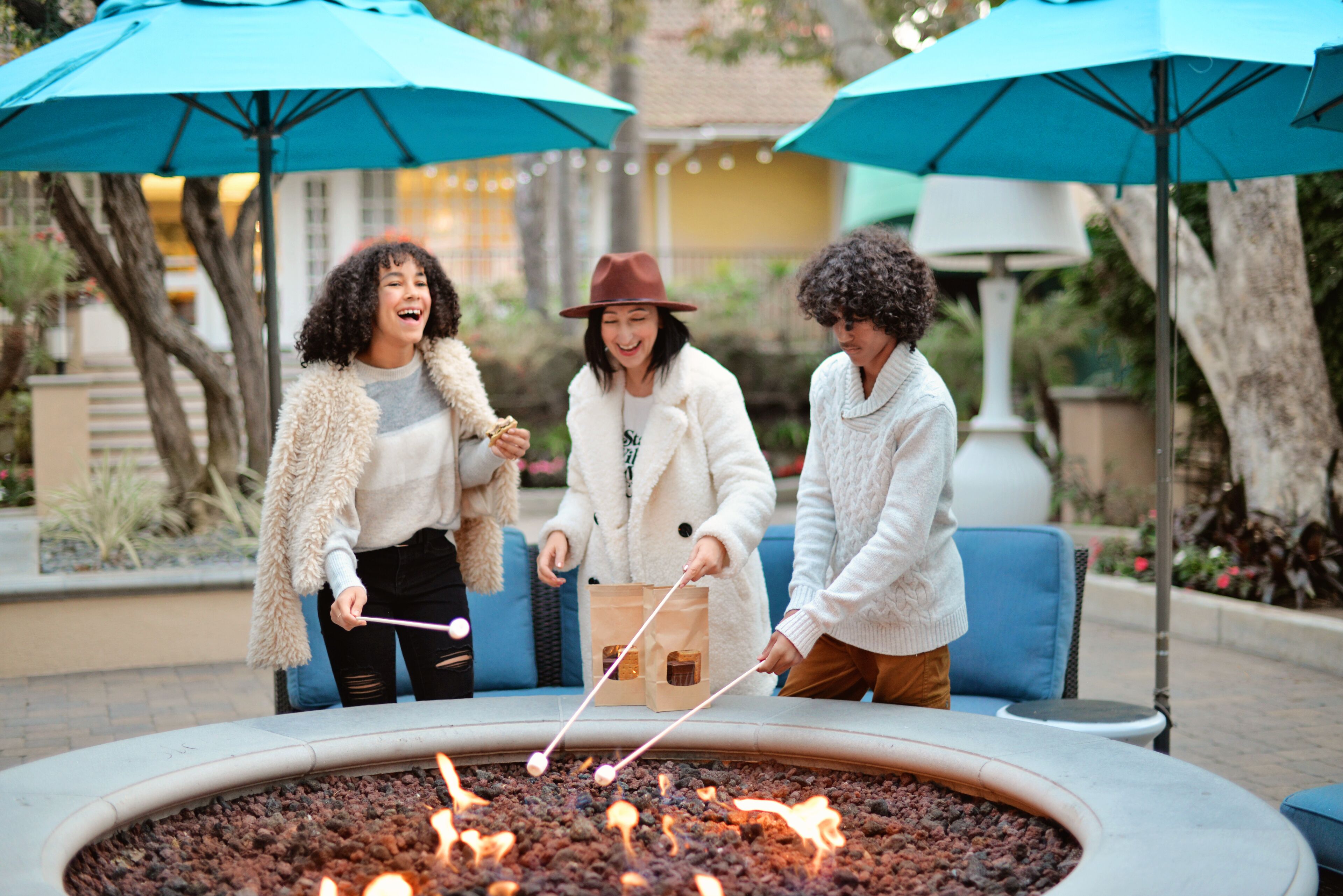Three people roasting marshmallows around a circular fire pit with blue umbrellas.