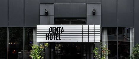 Una entrada de hotel moderna con un letrero que dice "OENTA HOTEL". Hay áreas de estar al aire libre con sillas y plantas en macetas.