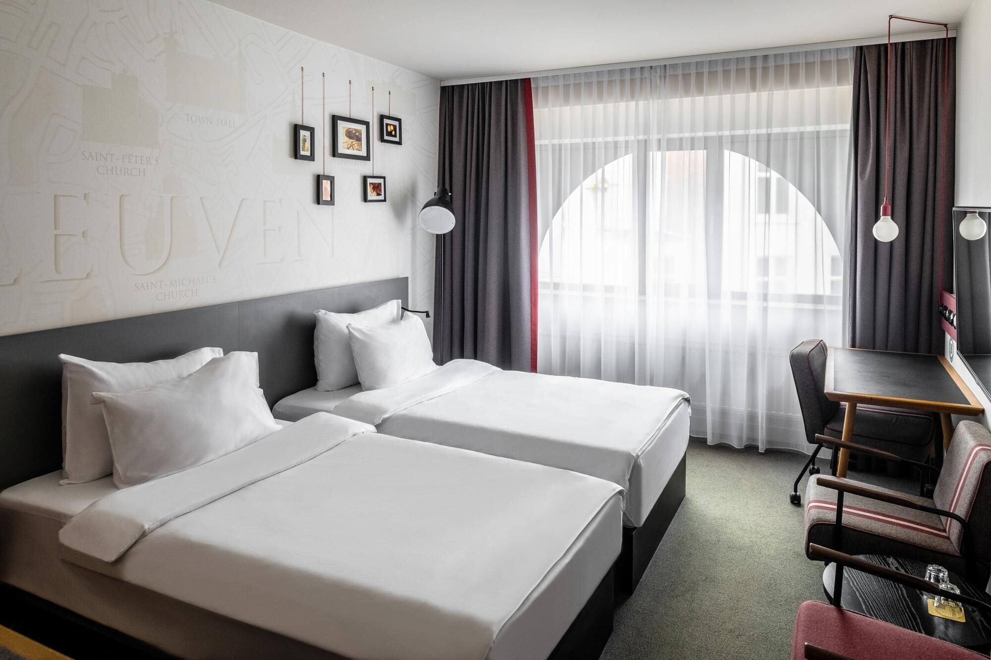 A hotel room with two beds, a desk, and a chair. There are framed pictures on the wall and a map.