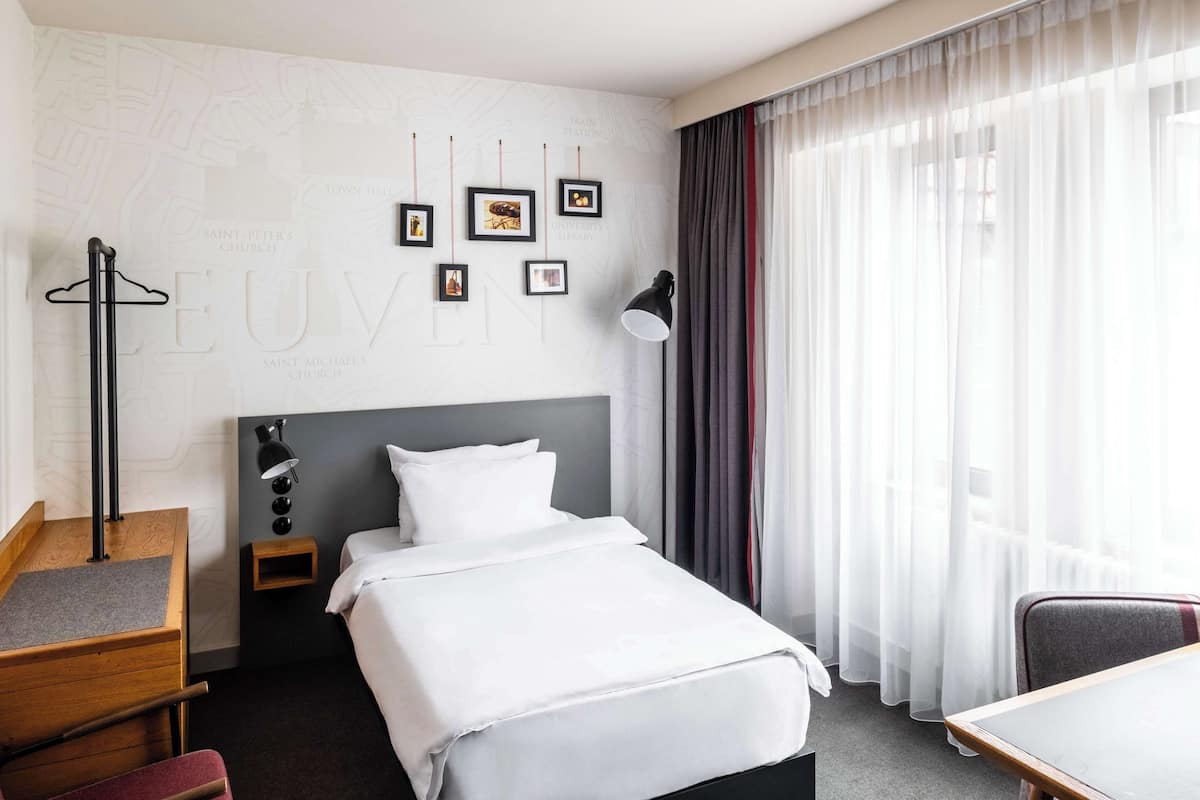 A hotel room with a bed, a desk, a chair, a nightstand, a lamp, and a window with curtains.