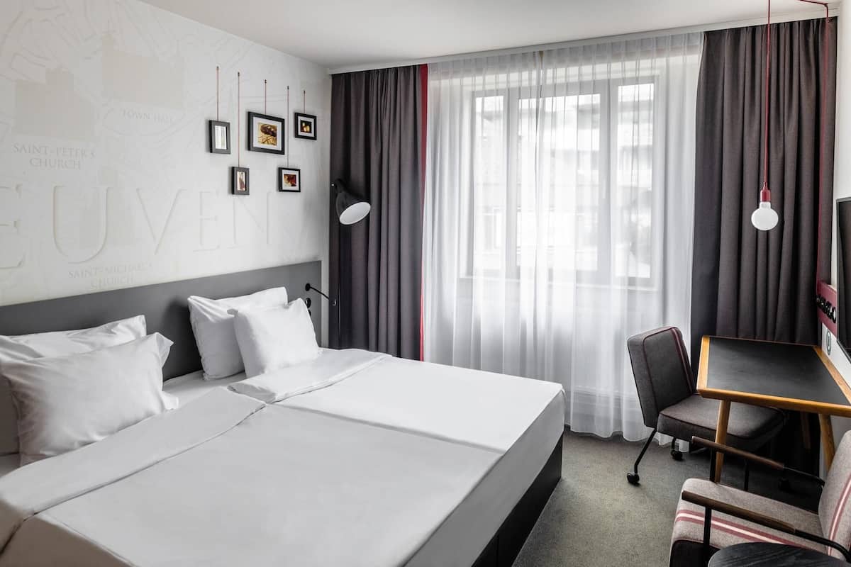 A hotel room with a large bed, a desk, and a chair. There are curtains on the windows, and a lamp is on the desk.
