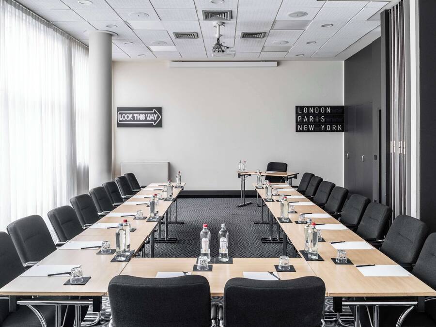 Una sala de conferencias con una larga mesa, sillas, botellas de agua y un cartel con indicaciones a Londres, París y Nueva York.