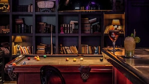 Una mesa de billar con bolas, una vara de billar y un vaso de bebida. Se ven estanterías llenas de libros y un reloj en la pared.