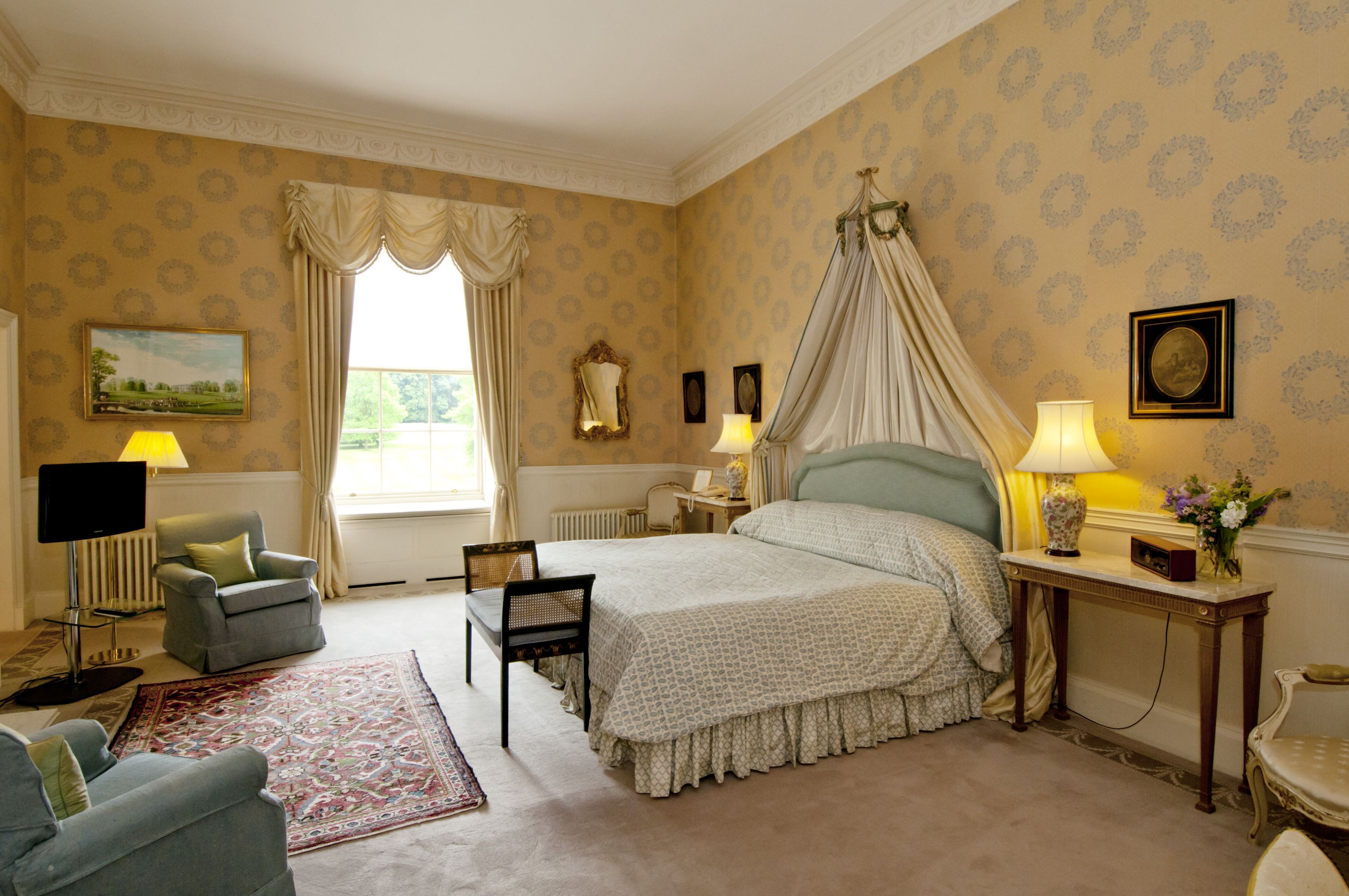 A bedroom with a four-poster bed, a sitting area with a chair and a table, a television, and a window with curtains.