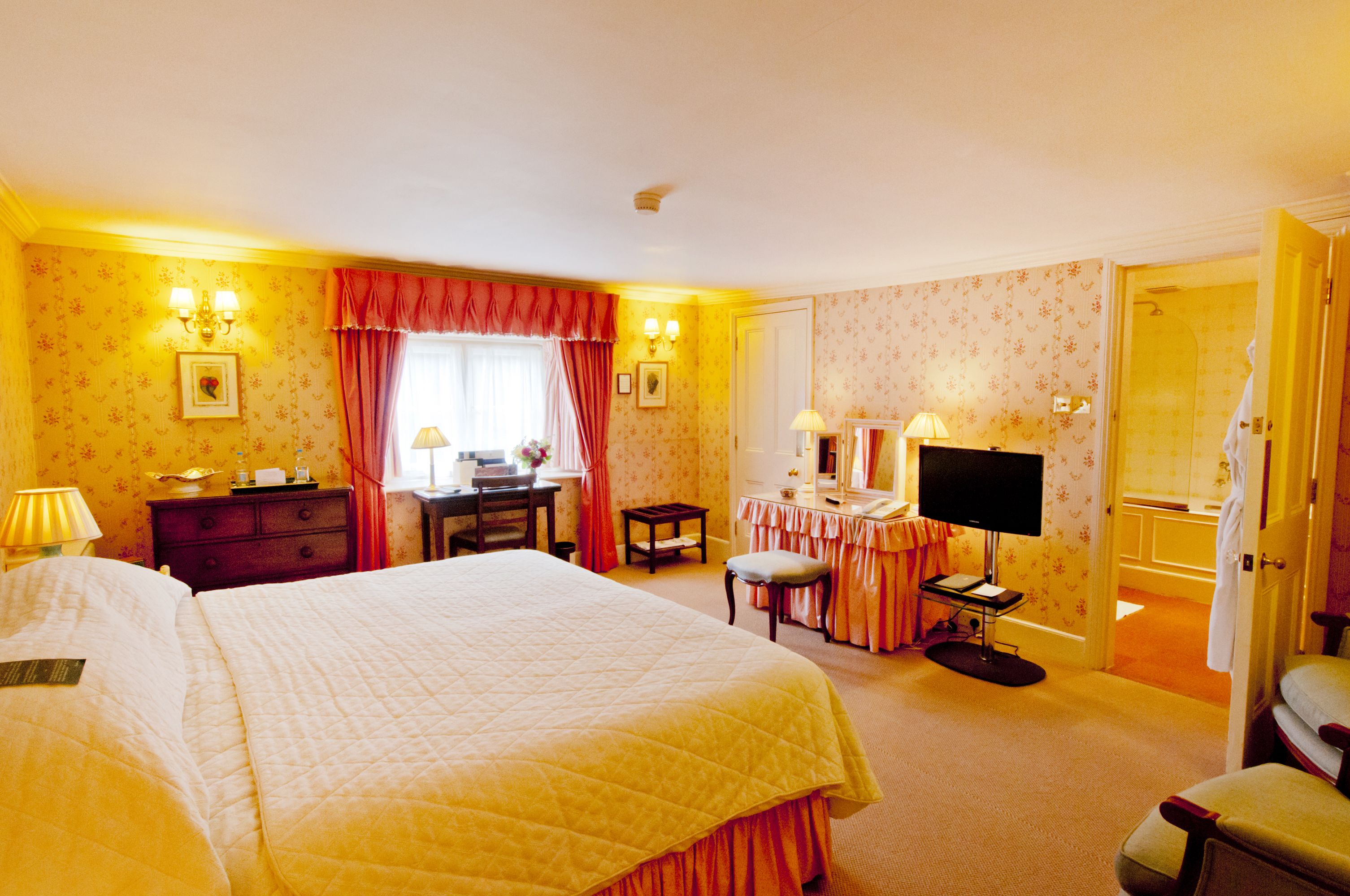 A hotel room with a large bed, a desk with a lamp, a vanity with a mirror, and a television.