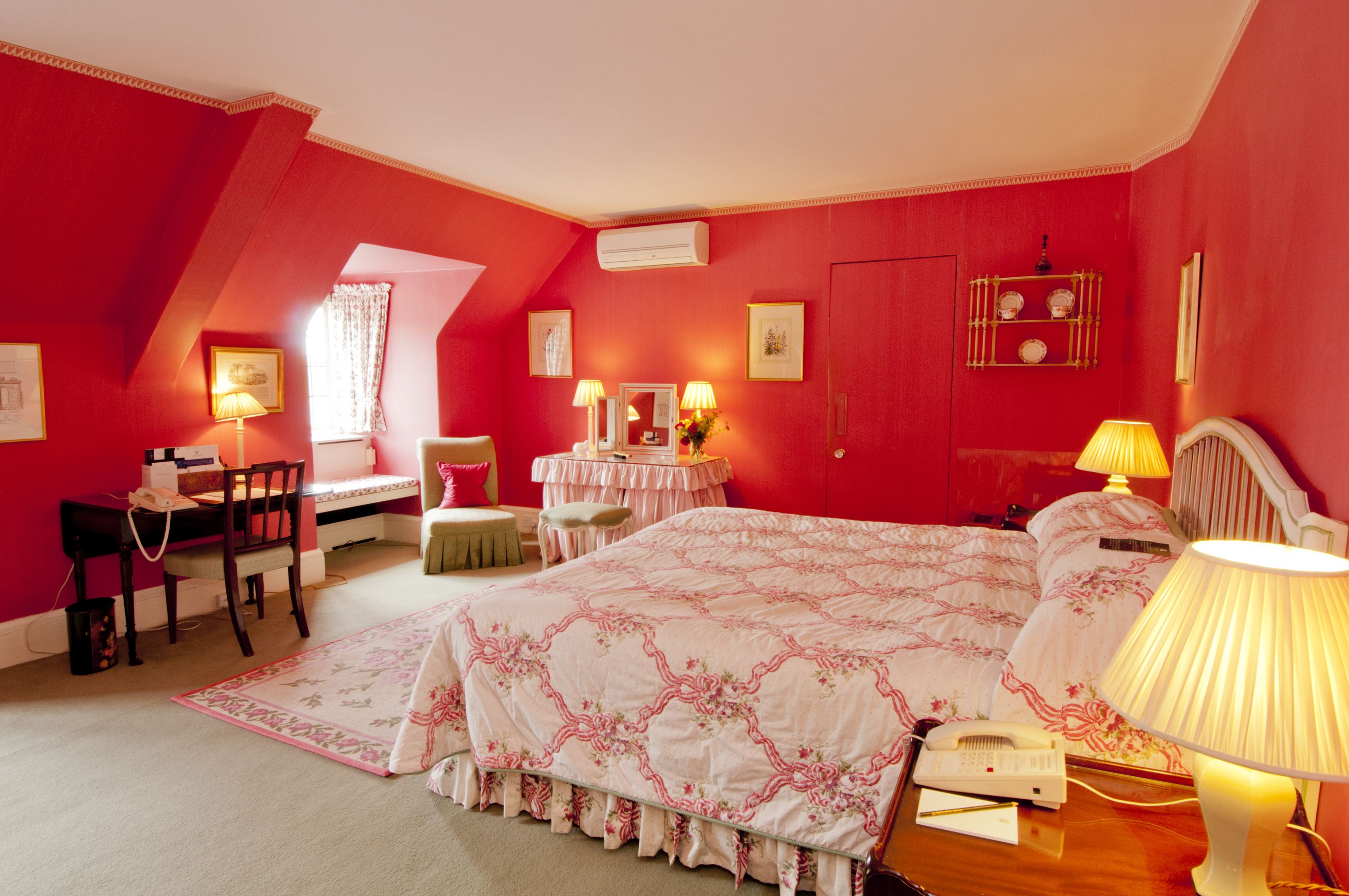 A bedroom with a sloped ceiling, a bed with a floral bedspread, two bedside tables with lamps, a desk with a chair, and a window with curtains.
