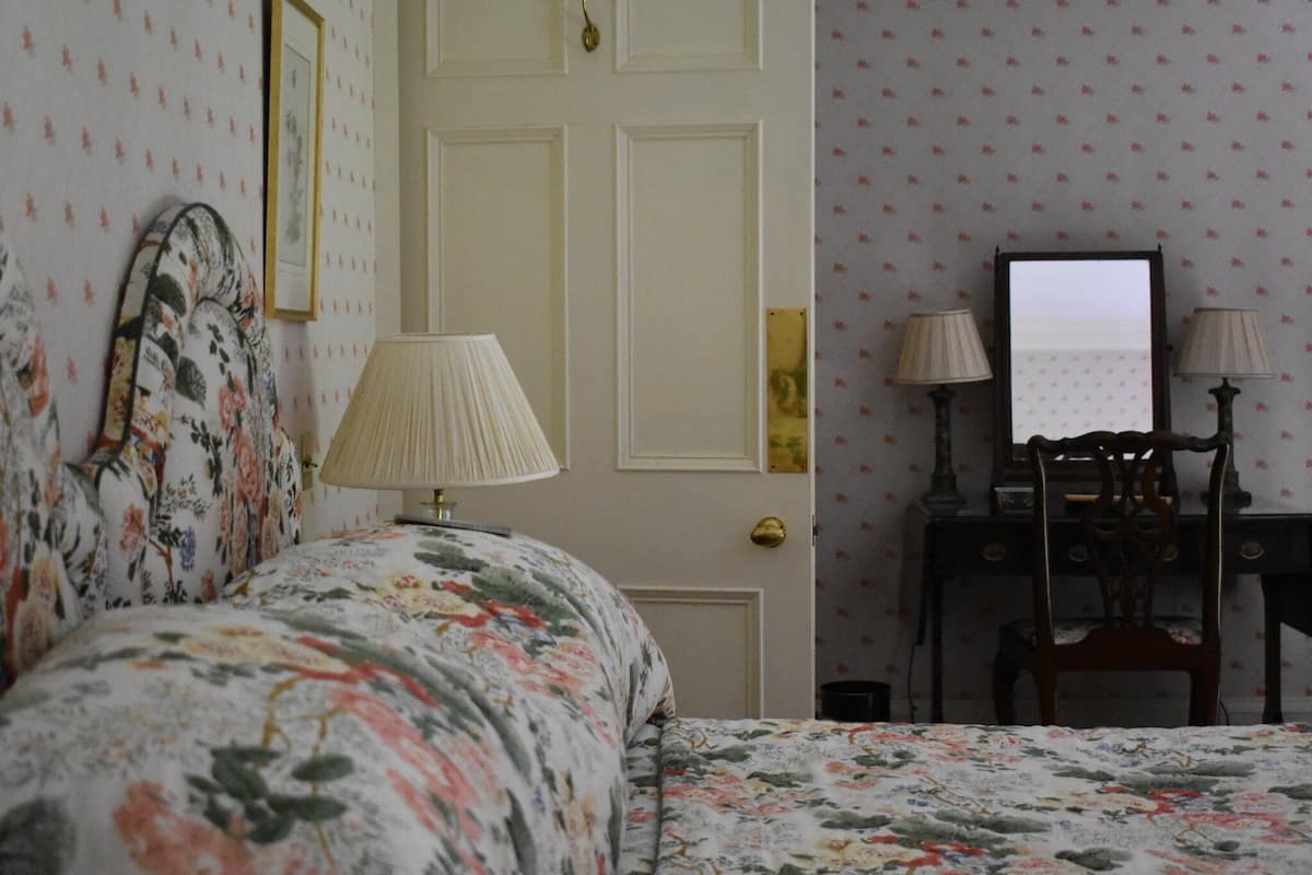 A bedroom with a floral bedspread, a bedside lamp, a dresser with a mirror, and a door with a brass handle.
