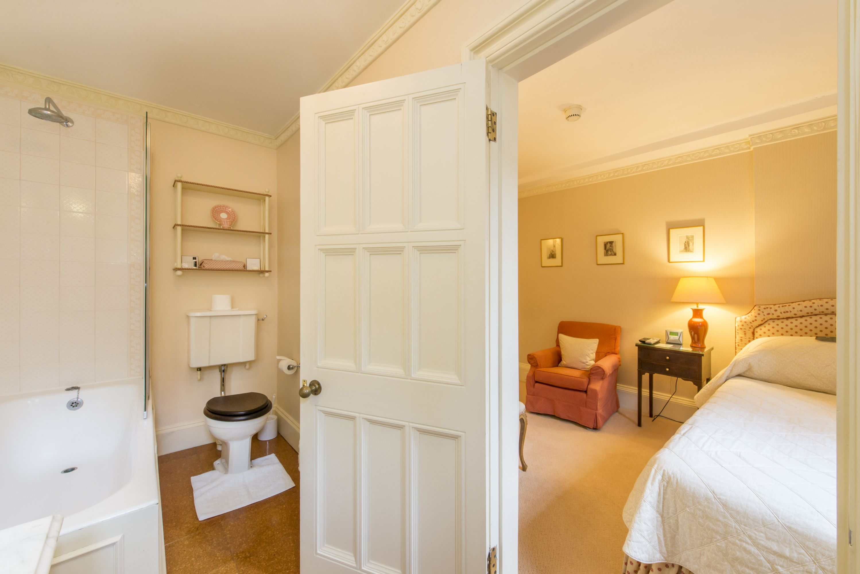 A room with a white door, a bathroom with a toilet and sink, a bed, a chair, and a nightstand with a lamp.