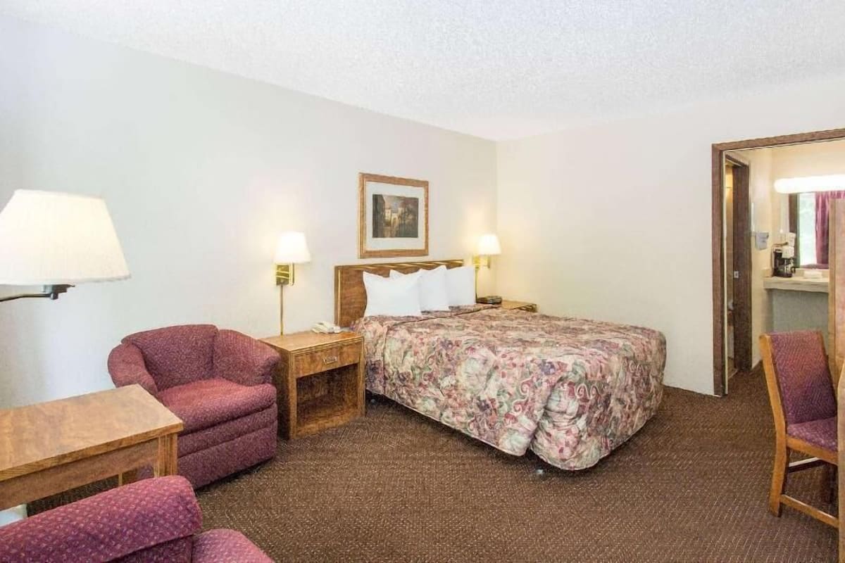 A hotel room with a bed, two chairs, a nightstand, a lamp, and a framed picture on the wall.
