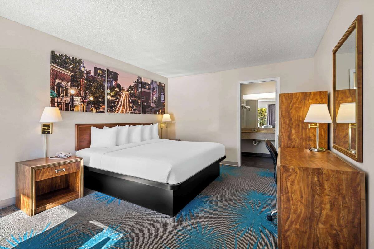A hotel room with a large bed, a nightstand, a desk with a mirror, and a cityscape painting on the wall.