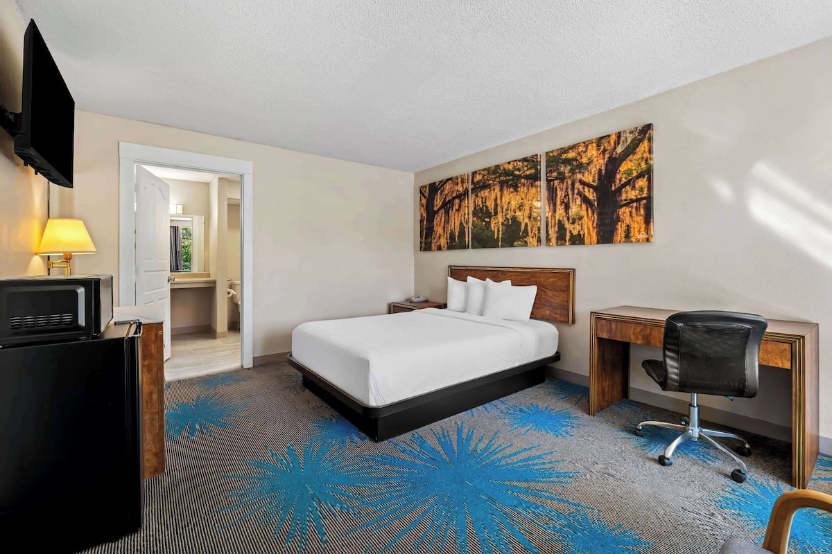 A hotel room with a bed, desk, chair, TV, and a painting on the wall.