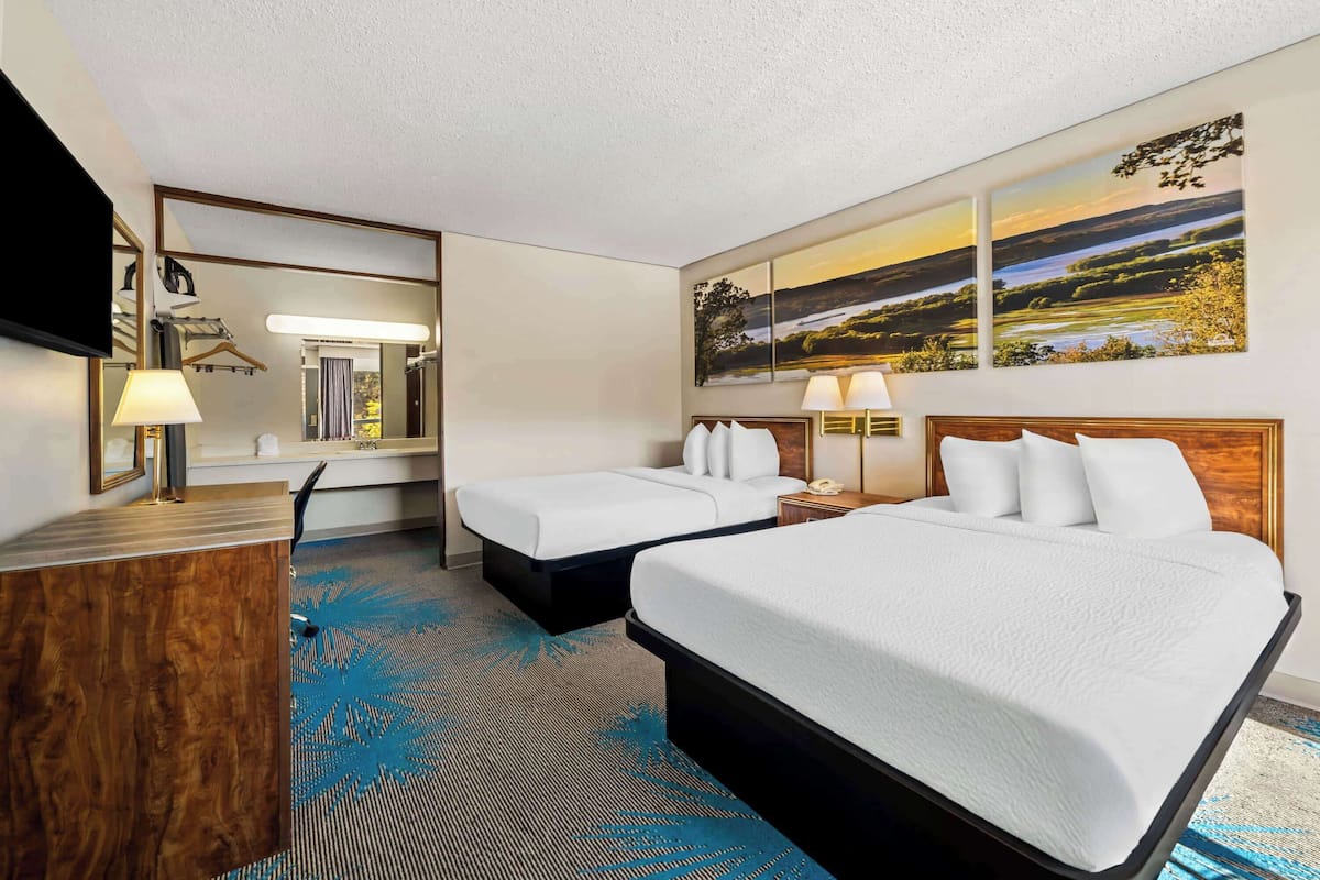 A hotel room with two beds, a desk, a mirror, and artwork on the wall.
