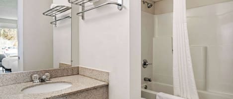 Un baño con lavamanos de encimera de granito, un espejo, ducha con cortina blanca y bañera.