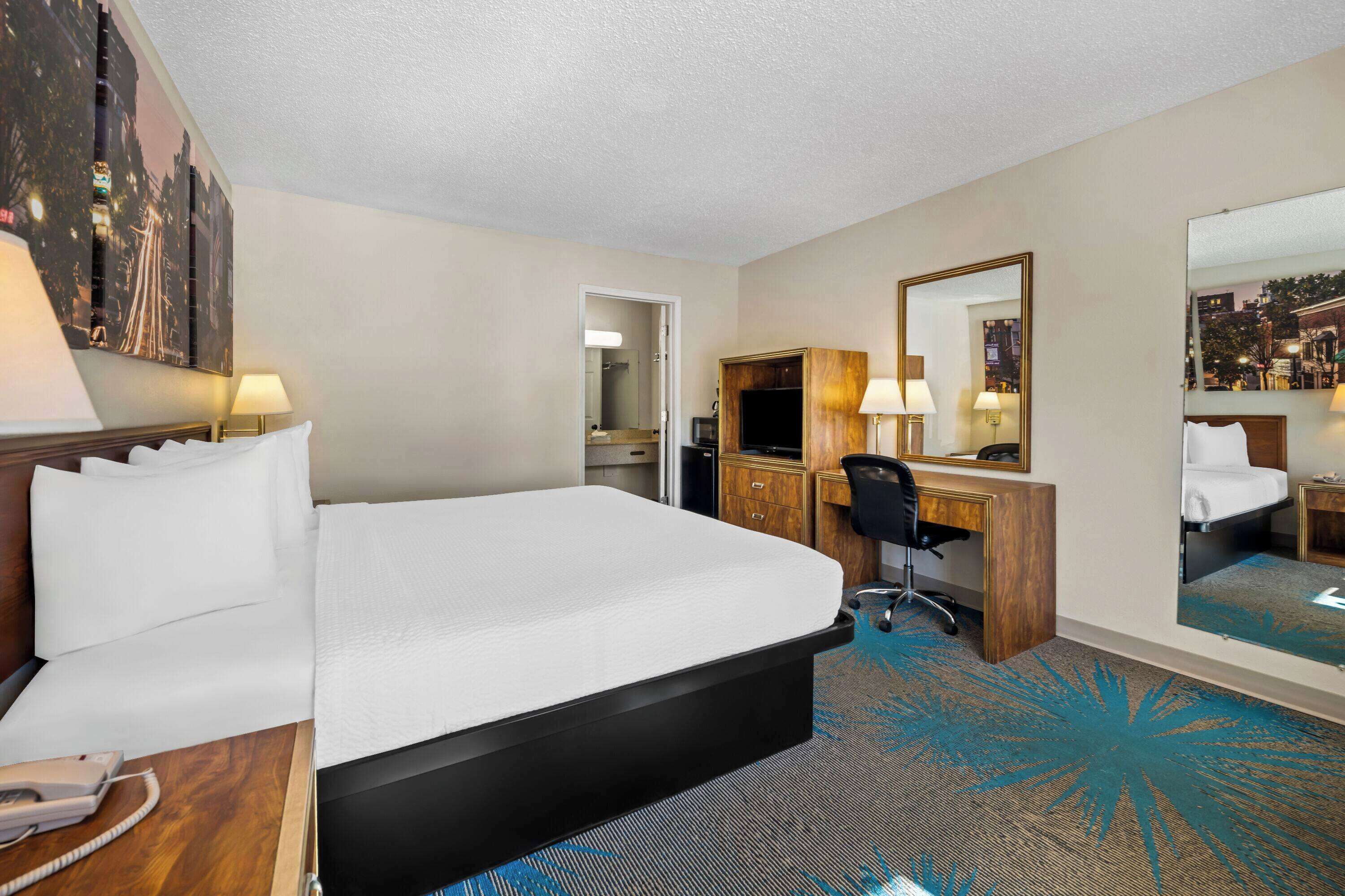 A hotel room with a large bed, a desk, a chair, a mirror, and a carpet with a blue pattern.