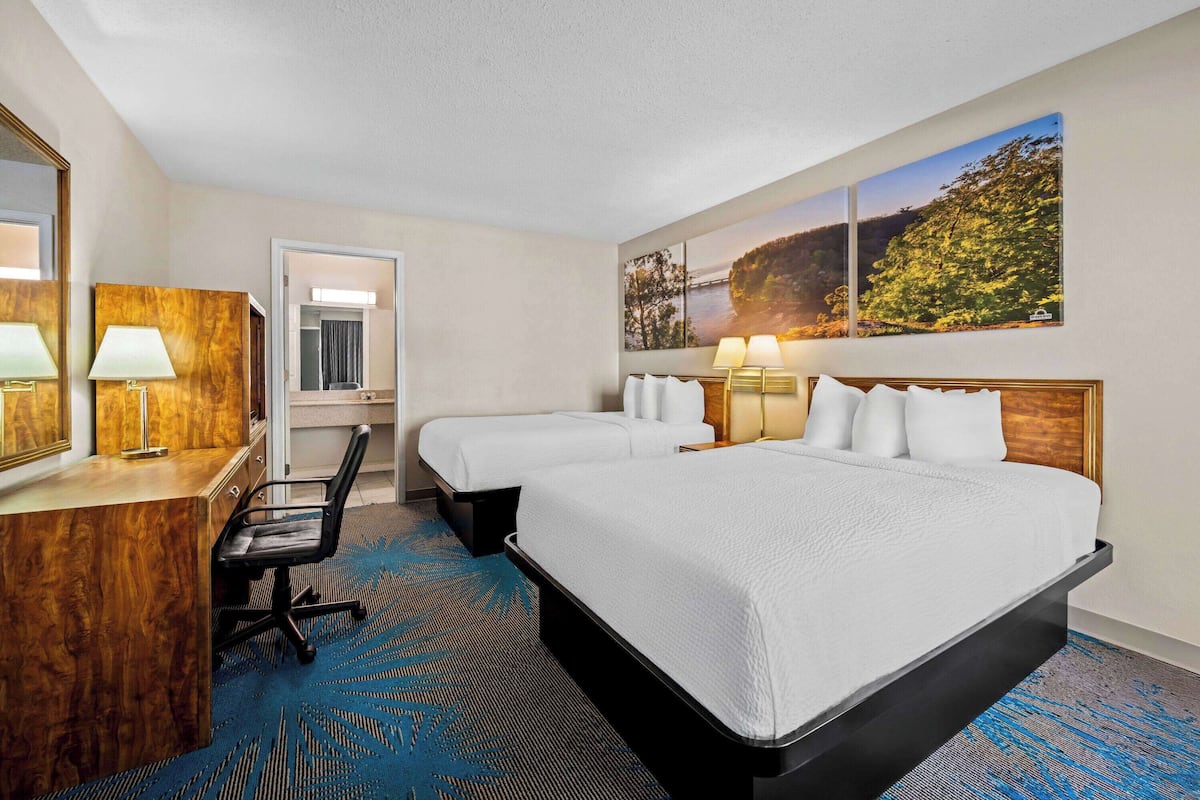 A hotel room with two beds, a desk with a chair, a desk lamp, a mirror, and a large window with a view of trees.