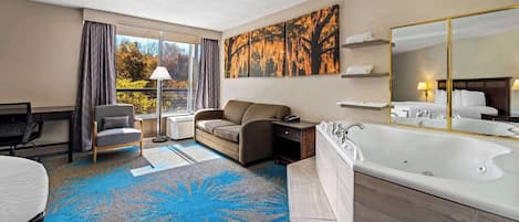 A modern hotel room with a large window, a sofa, a desk, a chair, a large bathtub, and a painting on the wall.