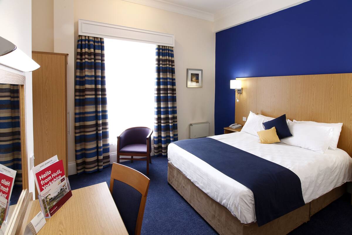 A hotel room with a bed, a chair, a table, and a window with curtains.