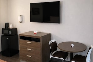 Habitación de hotel con televisión de pantalla plana en la pared, un escritorio de madera con cajones, un refrigerador negro compacto, un microondas, dos sillas marrones alrededor de una mesa redonda y un accesorio de luz blanco montado en la pared.