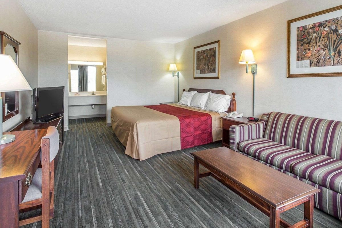 A hotel room with a bed, a sofa, a desk with a TV, a lamp, and a painting on the wall.
