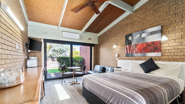 A modern hotel room with a brick wall, a large bed with a striped bedspread, a ceiling fan, a flat-screen TV, and a dining area with a wooden table and chairs. - Baronga Motor Inn (Colac)