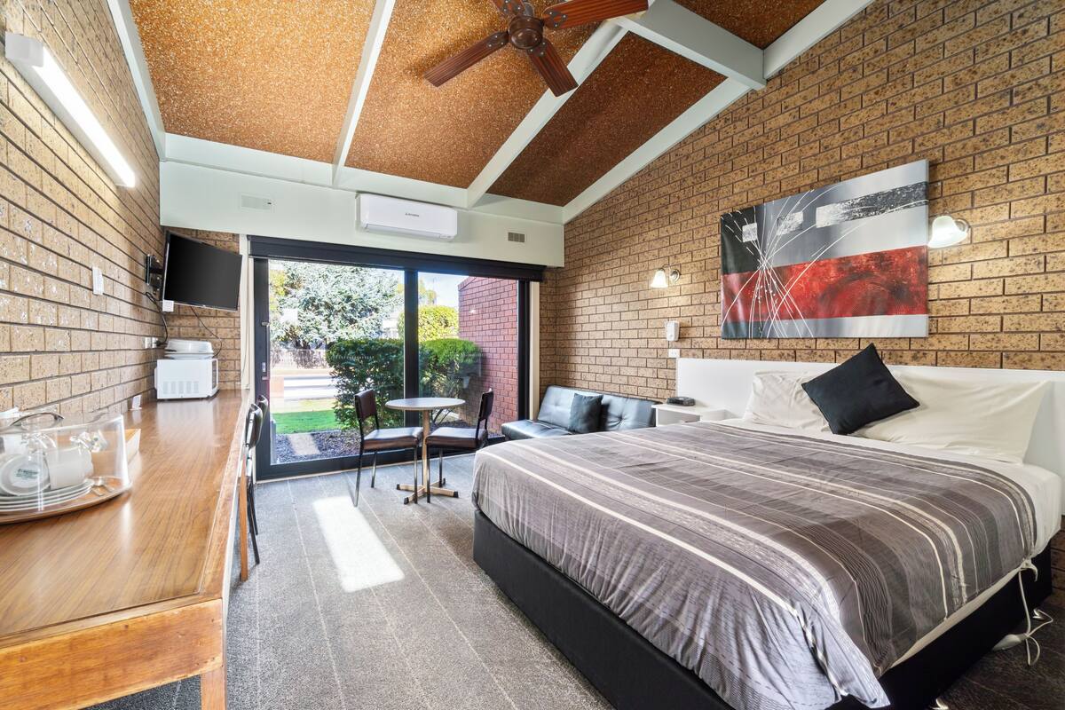 A modern hotel room with a brick wall, a large bed with a striped bedspread, a ceiling fan, a flat-screen TV, and a dining area with a wooden table and chairs.