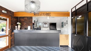 A reception area with a 'WELCOME' sign, a brick wall, a wooden ceiling, a tiled floor, and a black door with a diamond pattern. - Baronga Motor Inn (Colac)