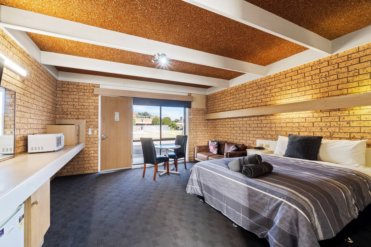A room with a brick wall, a bed with a striped bedspread, a desk with a microwave, and a chair.
