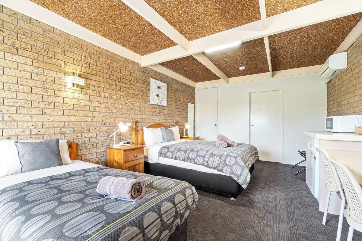 A hotel room with two beds, a desk, a chair, a microwave, and a brick wall.