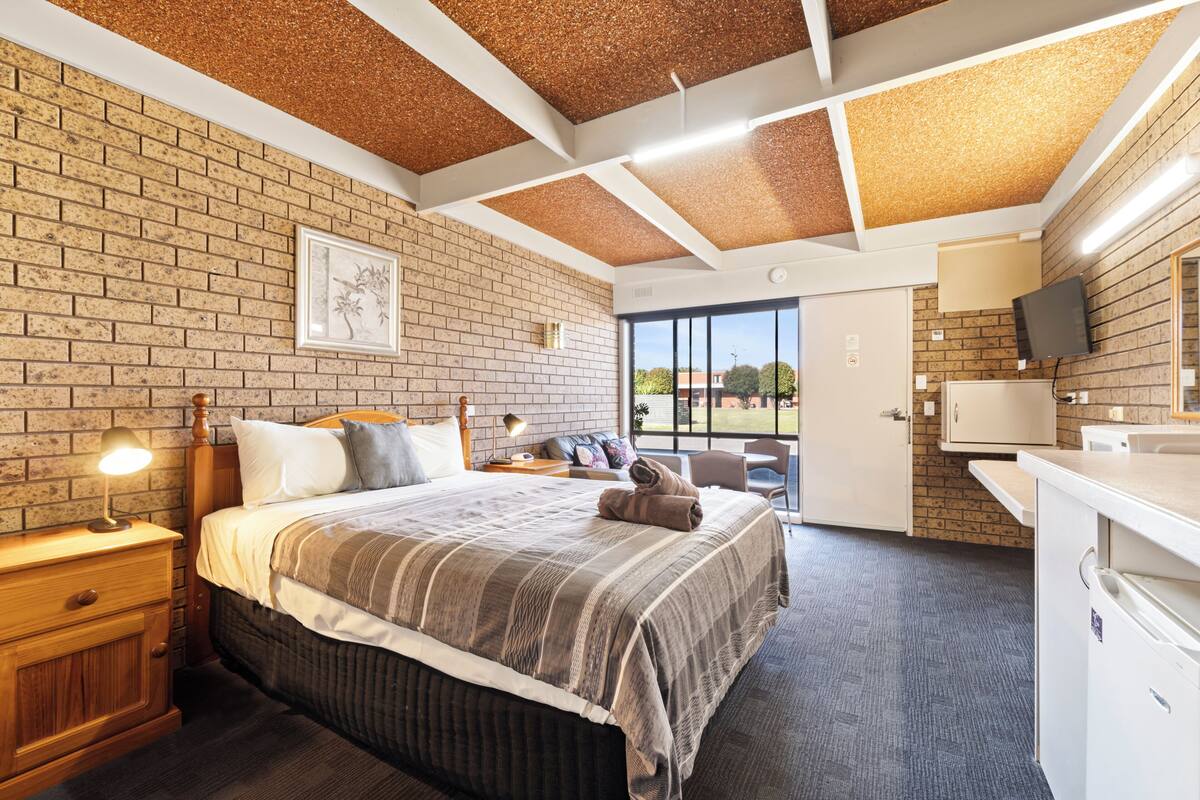 A hotel room with a brick wall, a bed with a striped blanket, a wooden nightstand, a desk, and a television.