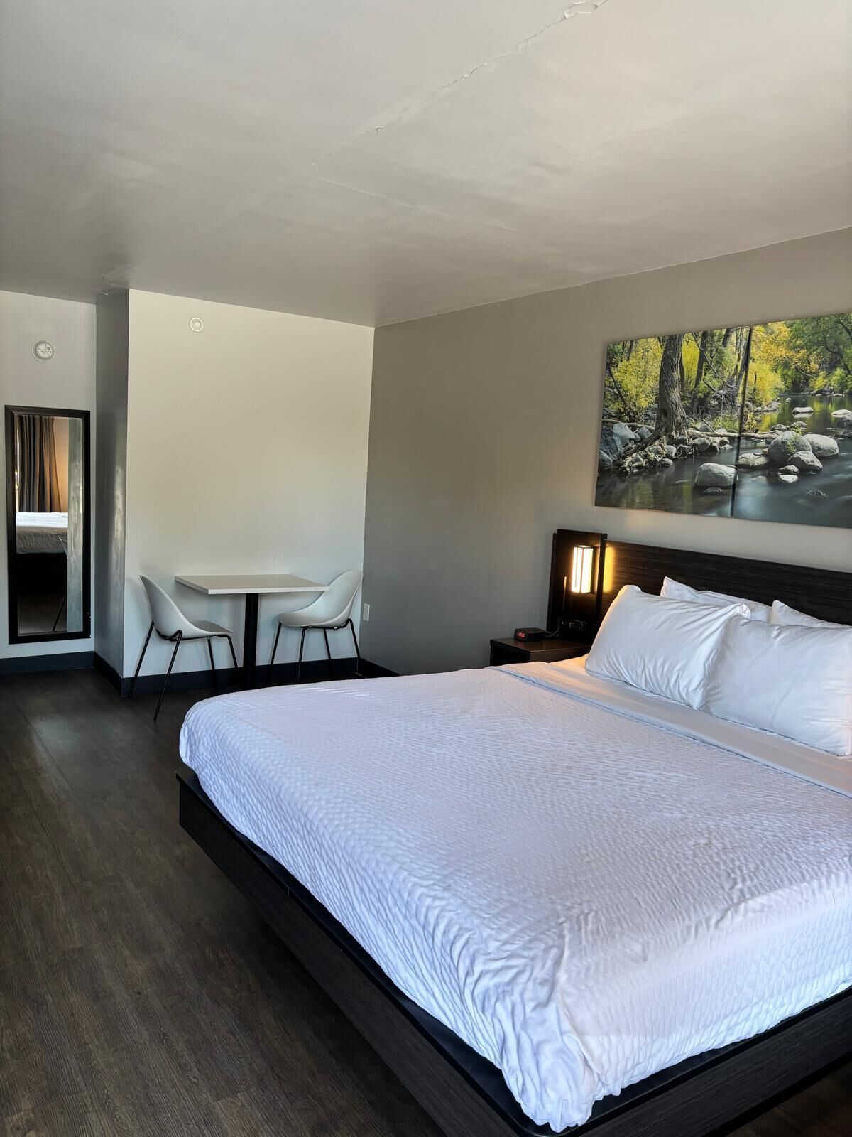 A modern hotel room with a large bed, a desk with a chair, and a painting of a forest scene.