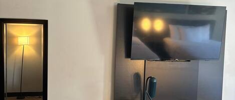 A hotel room with a bed, a desk with a chair, a television mounted on the wall, and a mirror.