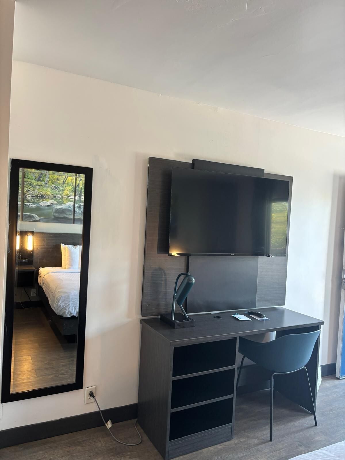 A modern hotel room with a bed, a desk with a chair, a flat-screen TV mounted on the wall, and a large mirror reflecting the room.