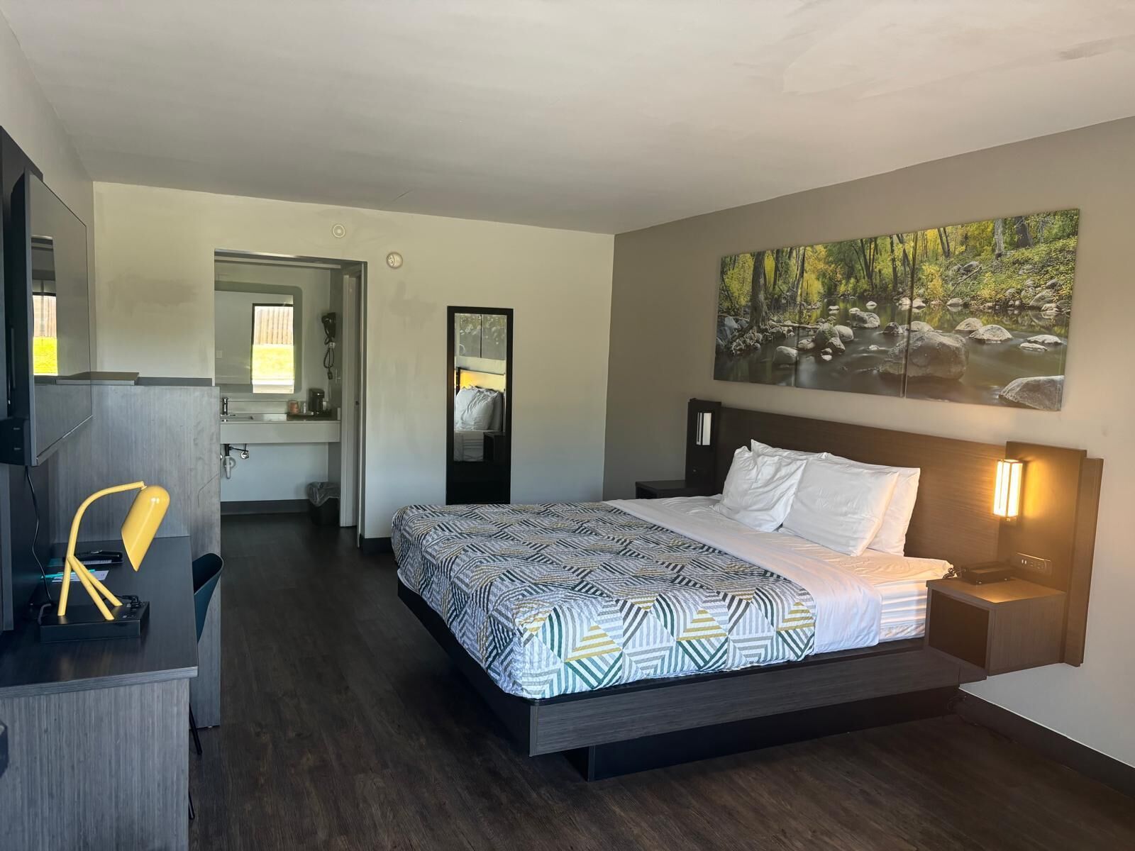 A hotel room with a bed, bedside table, a mirror, a desk with a lamp, a bathroom with a sink, and a painting on the wall.