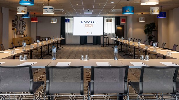 Een congreszaal met een lange tafel, stoelen en een projectorscherm waarop 'NOVOTEL' staat.
