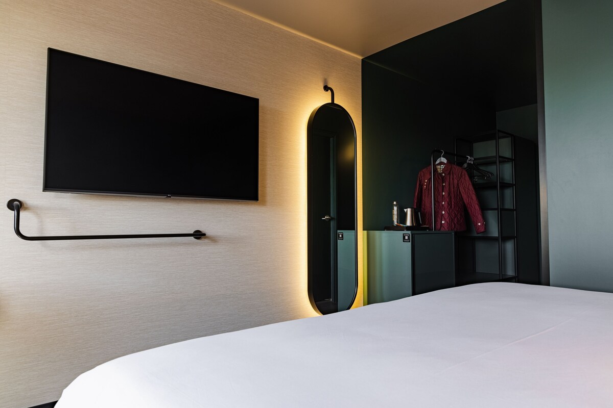 A modern hotel room with a bed, a flat-screen TV mounted on the wall, a coat rack with a red garment, and a black wardrobe.